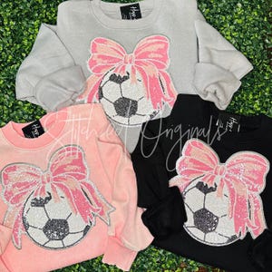 May include: Three sweatshirts with a pink and white bow and a soccer ball design. The sweatshirts are in grey, pink, and black.