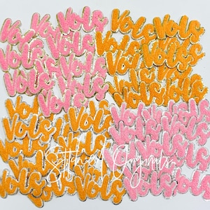 May include: A large pile of pink and orange felt patches with the word "Vote" written on them in a cursive font. The patches have a silver outline and are covered in glitter.