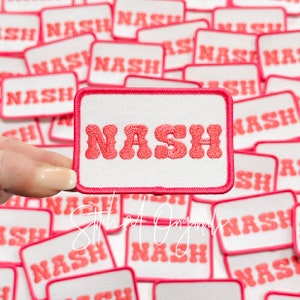 May include: A pink and white embroidered patch with the word "NASH" in a retro font. The patch is surrounded by a pink border.