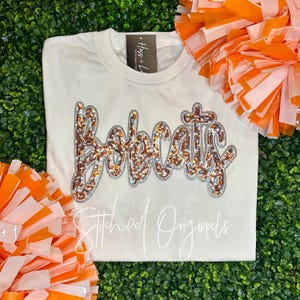 May include: Cream-colored sweatshirt with the word "Bobcats" in sequined lettering. The word is outlined in silver and filled with orange and brown sequins. The text "Stitched Originals" is written in cursive below the word. Orange and white pom-poms are in the background.