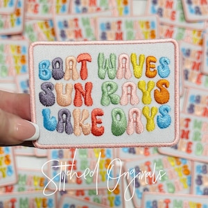 May include: Embroidered patch with the words "Boat Waves Sun Rays Lake Days" in colorful bubble letters. The patch has a white background with a pink border. The text "Stitched Originals" is written in cursive.