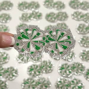 May include: Two silver glitter iron-on patches with green and white pom-poms. The patches are shaped like a circle and have a sparkly finish.