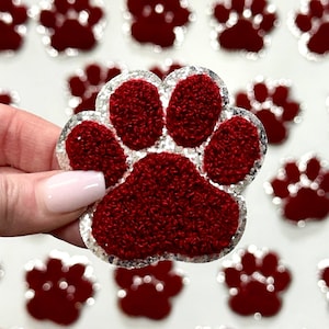 May include: Red glitter paw print iron-on patch with a white outline. The paw print is made of a fuzzy material.