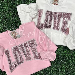 May include: Two pink and white sweatshirts with the word "LOVE" in large, sparkly pink letters. The sweatshirts are on a green background.
