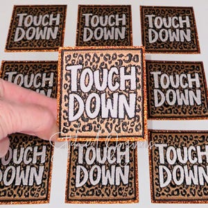 May include: Square patches with the words "TOUCH DOWN" in white block letters. The patches have a leopard print background and a glittery orange border. The patches are likely for sports apparel.