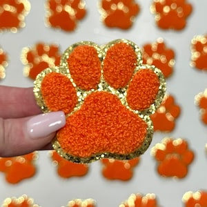 May include: Orange and gold glitter paw print iron-on patch. The paw print is fuzzy and has a gold glitter outline.
