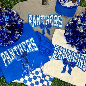 May include: A collection of apparel and accessories in blue, white, and gray, featuring the word "PANTHERS." Items include a blue baseball cap, t-shirts, a sweatshirt, and pom-poms. The word "PANTHERS" is in white with a blue outline and a bow.