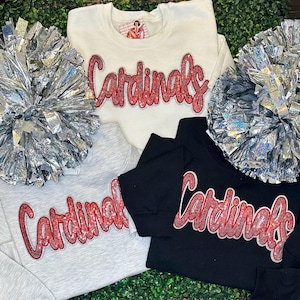 May include: A collection of sweatshirts in white, gray, and black, each emblazoned with the word "Cardinals" in red sequined lettering. Silver pom-poms are placed around the sweatshirts. The sweatshirts are likely team apparel.