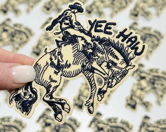 May include: A black and white embroidered patch of a cowboy riding a bucking horse with the text "Yee Haw" in a banner above the horse.