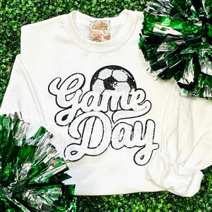 May include: White long-sleeved sweatshirt with a black and white sequined design of a soccer ball and the words "Game Day".