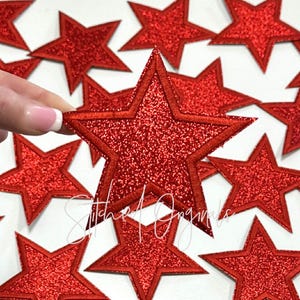 May include: A close-up of a pile of red glitter star-shaped iron-on patches. The stars have a double outline and are approximately 2 inches in size.