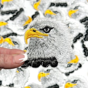 May include: Close-up of an embroidered iron-on patch featuring a white bald eagle head with a yellow beak and black eyes. The eagle's head is facing the right side of the image.