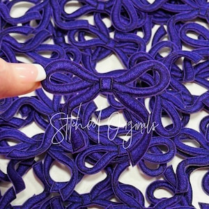 May include: A close-up of a pile of purple fabric bows. The bows are all the same size and shape, and they are arranged in a random pattern. The bows are made of a soft, velvety fabric and have a subtle shimmer.
