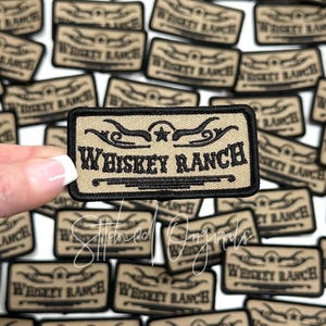 May include: A close-up of a tan embroidered patch with black stitching. The patch features a star and the text "WHISKEY RANCH".