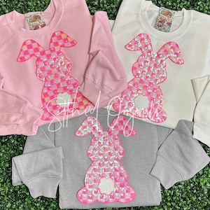 May include: Three sweatshirts with a pink and white checkered bunny design. The sweatshirts are in pink, white, and gray.