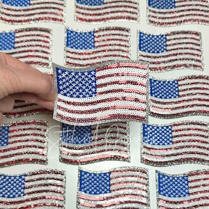 May include: A sheet of multiple sparkly American flag iron-on patches. The patches are made of sequins and have a silver glitter background. The flags are red, white, and blue.