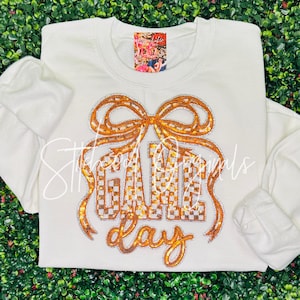 May include: White sweatshirt with a gold sequined bow and the words "GAME DAY" in gold sequins. The sweatshirt has long sleeves and a crew neck. The background is a green, leafy texture.
