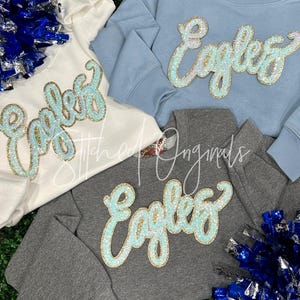 May include: Four sweatshirts in white, light blue, and gray, each with the word "Eagles" in a sequined, cursive font. The sweatshirts are arranged on a green surface with blue and silver pom-poms.