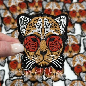 May include: A close-up of an embroidered iron-on patch featuring a cheetah wearing red sunglasses. The cheetah has a brown and white spotted pattern. The patch is surrounded by other cheetah patches.