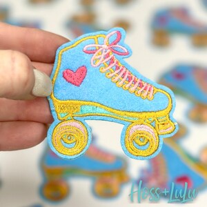 May include: A light blue roller skate patch with a pink heart and yellow and pink accents. The patch has yellow wheels and pink laces. The text "Hoss + Lulu" is in the bottom right corner.