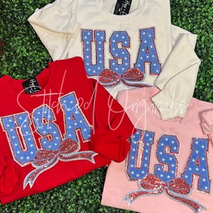 May include: Four sweatshirts in red, white, and pink, each featuring the word "USA" in blue letters filled with white stars. A red, white, and silver sequined bow is below the word. The sweatshirts are on a green background.