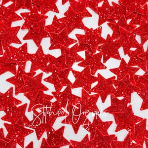 May include: A close-up image of a large quantity of red sequin star-shaped patches. The stars are scattered on a white surface.