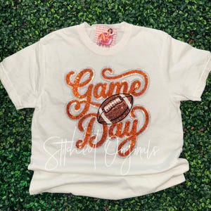 May include: A cream-colored short-sleeve t-shirt featuring a large, glittery orange and silver graphic that reads "Game Day" with a central football illustration. The design is centered on the chest, and a small brand tag is visible at the neckline.