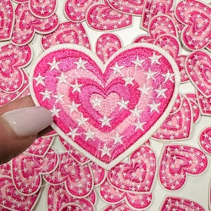 May include: A pink heart-shaped patch with white stars and a smaller pink heart in the center. The patch is surrounded by other pink heart-shaped patches with white stars.
