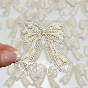 May include: A close-up of a white felt applique of a gold and white bow. The bow is a popular embellishment for clothing, accessories, and crafts.