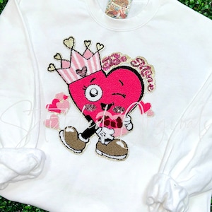 May include: White sweatshirt with a pink heart wearing a pink and white striped crown. The heart has a winking eye, a pink mouth, and the words "Be Mine" written on it.