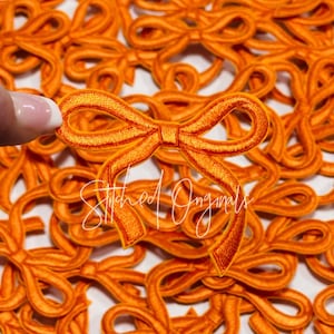 May include: A close-up of a pile of orange fabric bows. The bows are all the same size and shape, and they are arranged in a random pattern. The bows are made of a soft, velvety fabric and have a slightly textured surface.