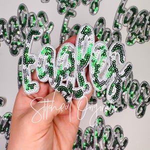 May include: A collection of sequined "Teacher" patches in a cursive font. The patches feature green and silver sequins with a white border. The patches are scattered on a white surface. The text "Stitched Originals" is also visible.