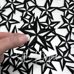 May include: Black and white embroidered star patches. The patches feature a five-pointed star design with a black outline and white center. The patches are approximately 2 inches in diameter. The image shows a hand holding one patch with many more in the background.