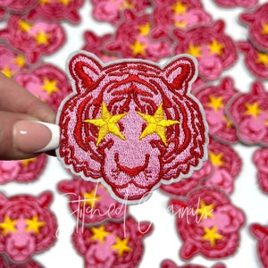 May include: Pink embroidered iron-on patch of a tiger with yellow star-shaped eyes. The patch is surrounded by other similar patches.