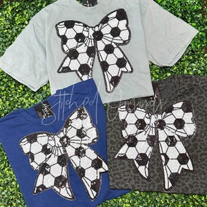 May include: Three t-shirts with a black and white soccer ball bow design. The shirts are in blue, gray, and black.