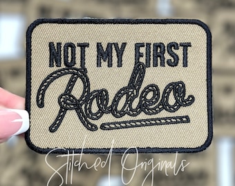 May include: Beige embroidered patch with black stitching. The patch features the text "NOT MY FIRST RODEO" with the word "RODEO" in a rope design.