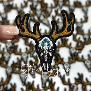 May include: Embroidered patch featuring a deer skull with antlers. The antlers are brown with a textured appearance, outlined in black. The skull is white with turquoise accents. The patch is held in front of a blurred background of similar patches. The text "Stitched Originals" is at the bottom.