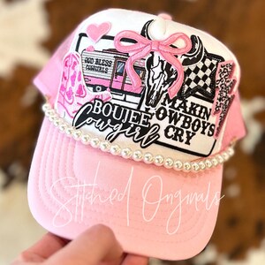 May include: Pink and white trucker hat with a variety of patches, including a pink bow, a skull, a camper with the text "God Bless Cowgirls", and a patch that says "Boujee Cowgirl Makin Cowboys Cry". The hat is decorated with a row of white pearls.
