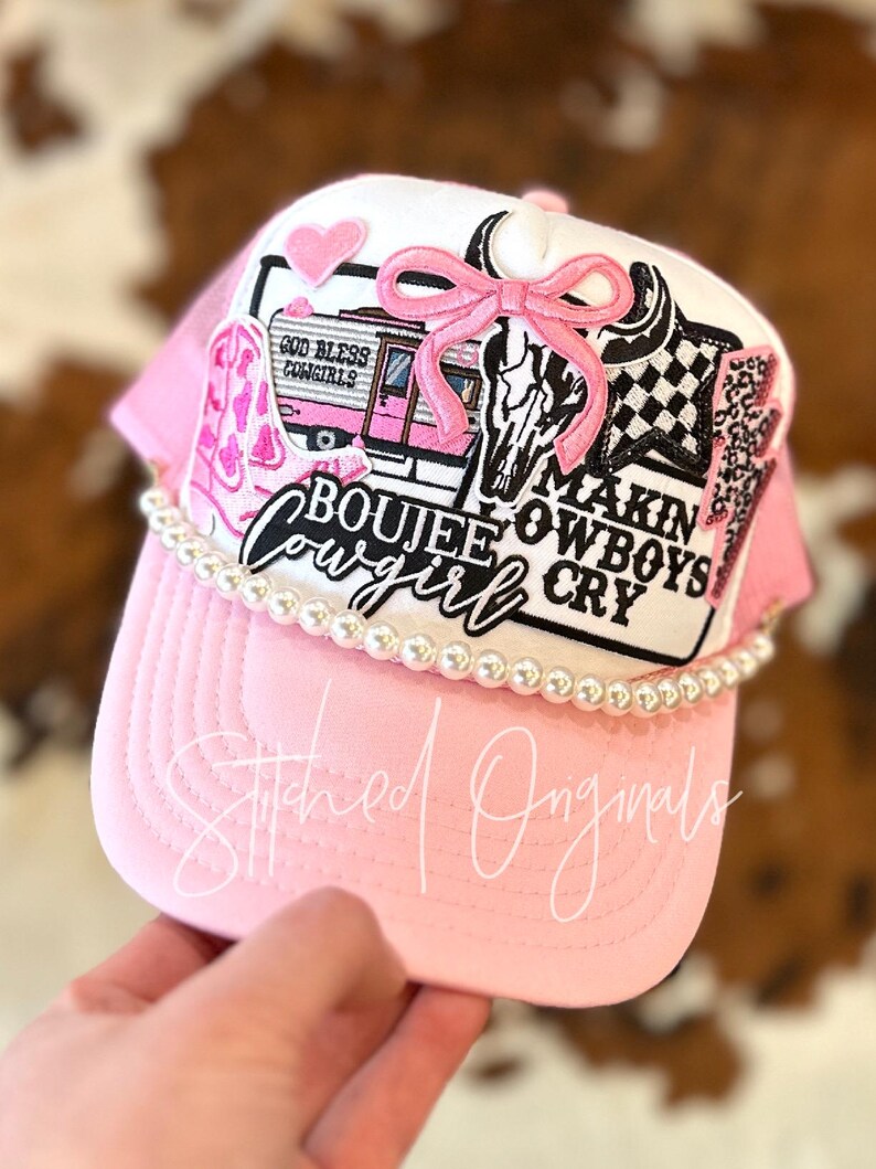 May include: Pink and white trucker hat with a variety of patches, including a pink bow, a skull, a camper with the text "God Bless Cowgirls", and a patch that says "Boujee Cowgirl Makin Cowboys Cry". The hat is decorated with a row of white pearls.