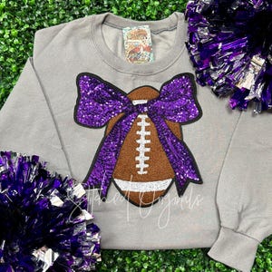 May include: A gray sweatshirt with a purple sequined bow and a brown football design. The football has white laces and a white outline.