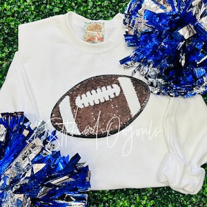 May include: White sweatshirt with a brown and white sequined football design. The sweatshirt has a crew neck and long sleeves.