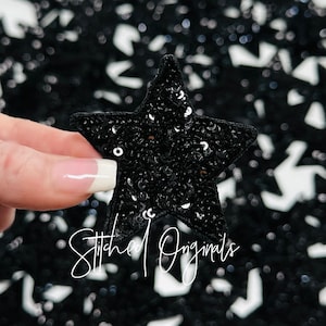 May include: A black sequined star-shaped patch with a white background. The patch is held in a hand and the text "Stitched Originals" is visible in the bottom of the image.