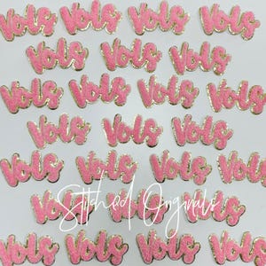 May include: Pink felt patches with gold glitter trim spelling out the word "Vols" in a cursive font.
