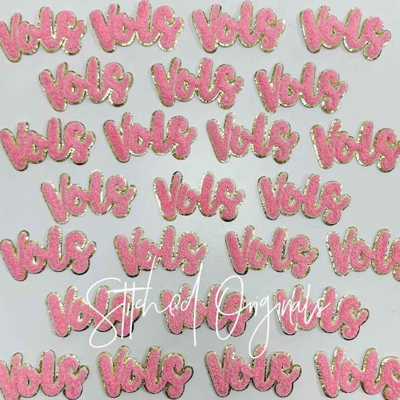 May include: Pink felt patches with gold glitter trim spelling out the word "Vols" in a cursive font.