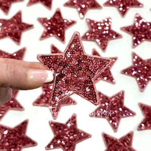 May include: A close-up of a pink sparkly star-shaped patch. The patch is made of sequins and has a pink satin border.
