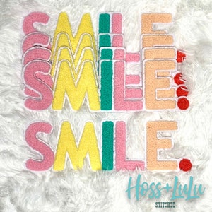 May include: A set of five colorful felt patches spelling out the word "SMILE". The patches are in shades of pink, yellow, green, and peach. The patches are arranged in a staggered pattern, with the word "SMILE" spelled out twice.