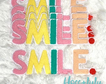 May include: A set of five colorful felt patches spelling out the word "SMILE". The patches are in shades of pink, yellow, green, and peach. The patches are arranged in a staggered pattern, with the word "SMILE" spelled out twice.