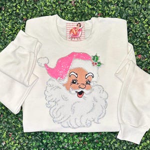 May include: White sweatshirt featuring a Santa Claus design with a pink sequined hat and a glittery white beard. The Santa face has a rosy complexion and a smiling expression. A small tag is visible near the neckline.