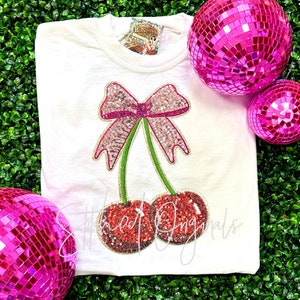 May include: White t-shirt with a pink sequined cherry design. The cherries have green stems and a pink bow. The shirt is on a green background with pink disco balls.