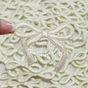 May include: A close-up of a white satin bow applique. The bow is made of a soft, delicate fabric and has a simple, elegant design.